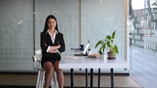 Mujer de negocios segura de sí misma sin traje negro sentada con los brazos cruzados en una oficina moderna.