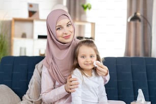 Familia árabe feliz, mamá musulmana y su pequeña hija linda, sentada en el sofá de casa y haciendo procedimientos de higiene matutinos. Madre limpiando la cara del niño con una servilleta de papel.