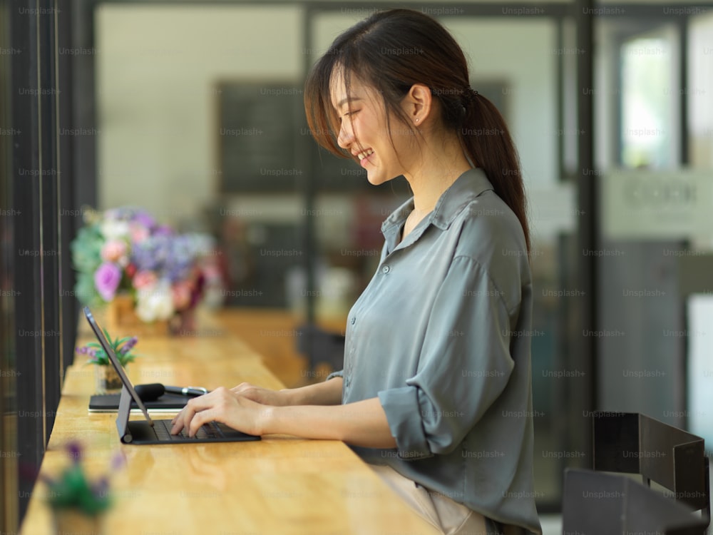Side view of young beautiful female smiling and working with digital tablet on wooden bar in coffee shop