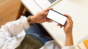 Top view of male freelancer hands holding horizontal smartphone with mock up screen above workspace, clipping path