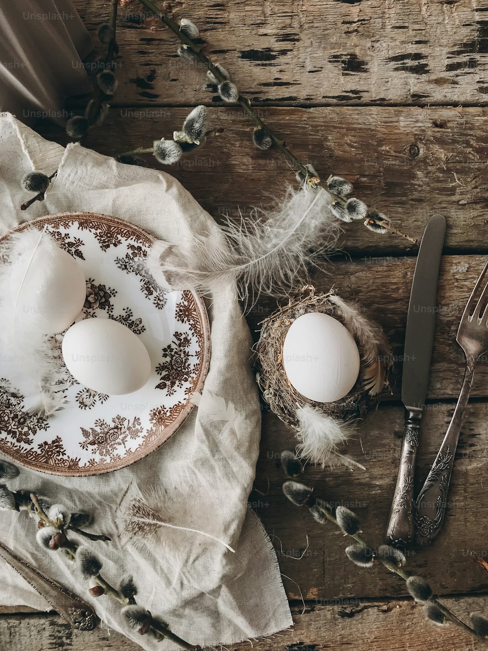 Stylish rustic Easter table setting flat lay. Natural easter eggs, feathers, vintage plate and cutlery, napkin, pussy willow branches on aged wooden table. Rural Easter still life. Happy Easter