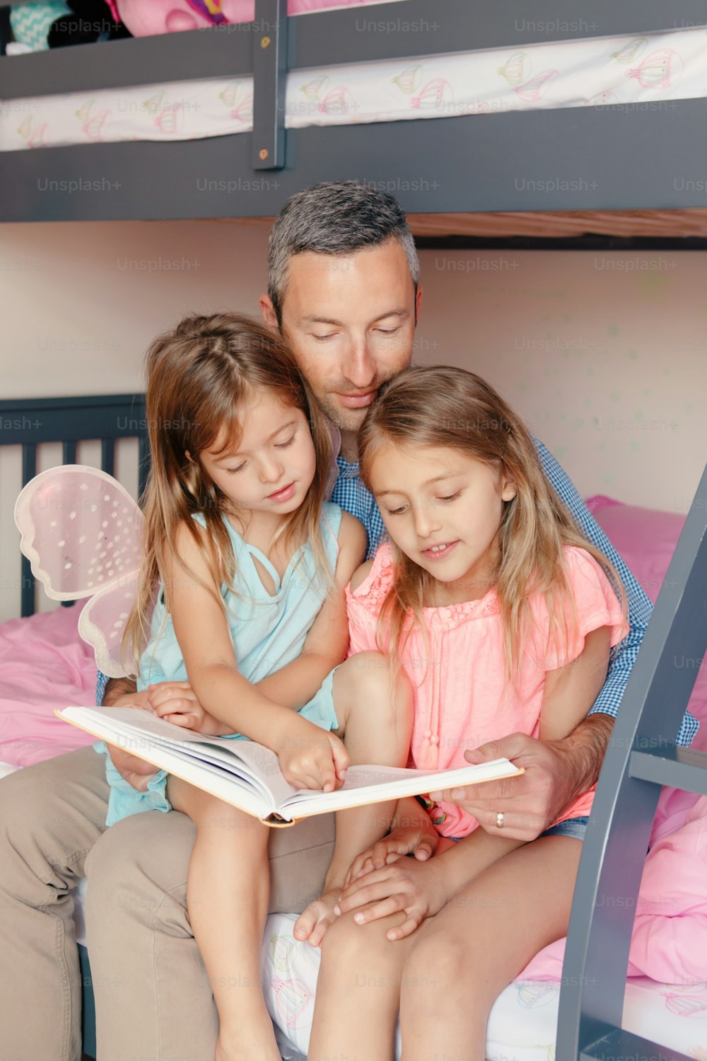 Fathers Day. Dad reading book to daughters girls. Family of three people sitting on bed in bedroom. Happy father and children at home spending time together. Happy lifestyle childhood.