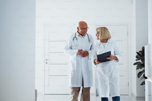 Two senior doctors stands in the clinic with notepad and talks to each other.