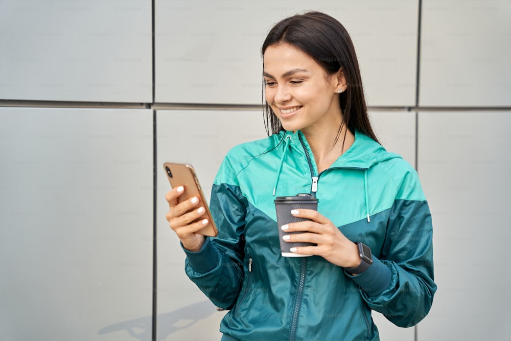 Waist up of smiling pretty lady holding coffee and looking at smartphone screen outdoor. Sport and lifestyle concept