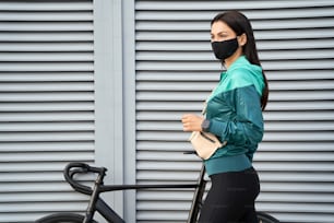 Woman in a sports jacket and leggings standing near a bicycle on the street and wearing face mask. Copy space