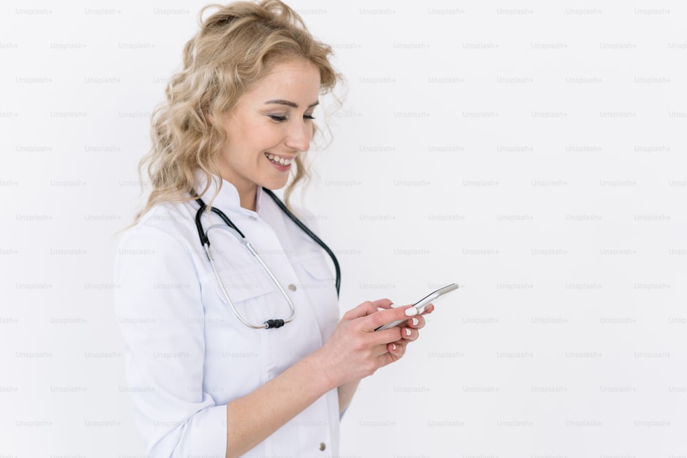 Aplicación móvil y concepto de tecnología moderna. Doctora feliz en uniforme médico blanco sostiene el teléfono inteligente, lee el mensaje del paciente, envía una consulta en línea de pie aislado en el fondo del espacio de copia