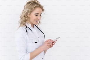 Mobile app and modern tech concept. Happy woman doctor in white medical uniform hold smartphone, reading message from patient, send online consultation standing isolated on copy space background