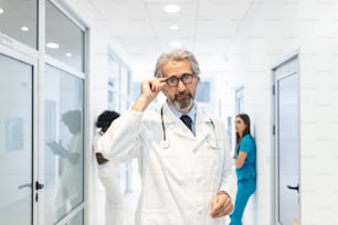 Portrait of confident mature doctor holding his glasses in hospital corridor. He is wearing lab coat in hospital.