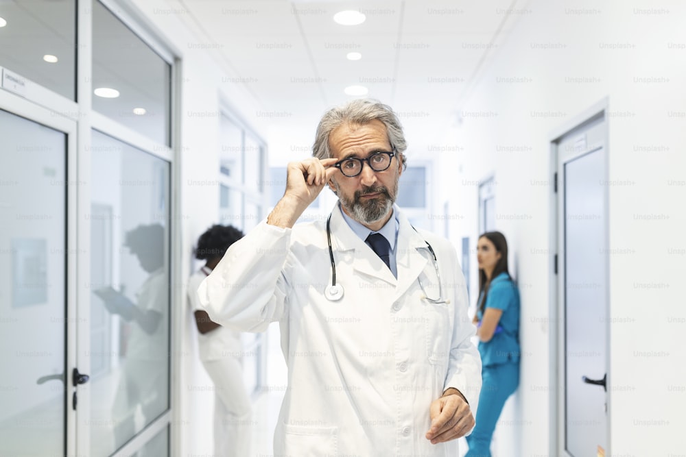 Porträt eines selbstbewussten reifen Arztes, der seine Brille im Krankenhausflur hält. Er trägt einen Laborkittel im Krankenhaus.