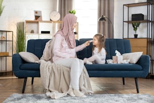 Young happy Arabian woman mother, wearing headscarf, wiping with napkin face of her cute little 3 years old daughter, sitting together on blue sofa at cozy room at home.