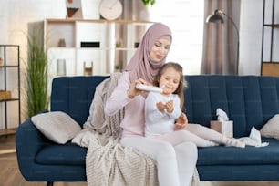 Skin moisturizer, hygiene concept. Young muslim mother in hijab sitting on the sofa at home with her cute smiling little daughter, holds bottle with body milk cream and pouring it on girl hands.