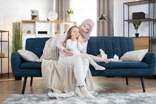 Happy Arabian mother sits on sofa at home and hugs her little daughter. Adorable smiling little girl after bath, with cream on her face, copying mom behavior, using cosmetic cream at home.