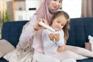 居心地の良いリビングルームで一緒に座っている間、彼女の小さな娘の手にスキンクリームやローションを塗る、ヘッドスカーフの若いアラビアのお母さんのクローズアップ。肌の保湿剤、衛生コンセプト