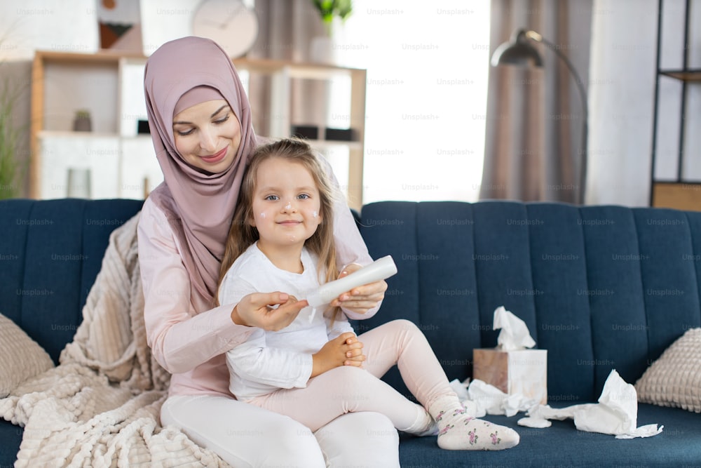 Bella giovane mamma musulmana in hijab, pronta ad applicare una crema o una lozione per la pelle sul viso della sua bambina, versando la crema da una bottiglia. Madre e figlia sedute sul divano a casa.