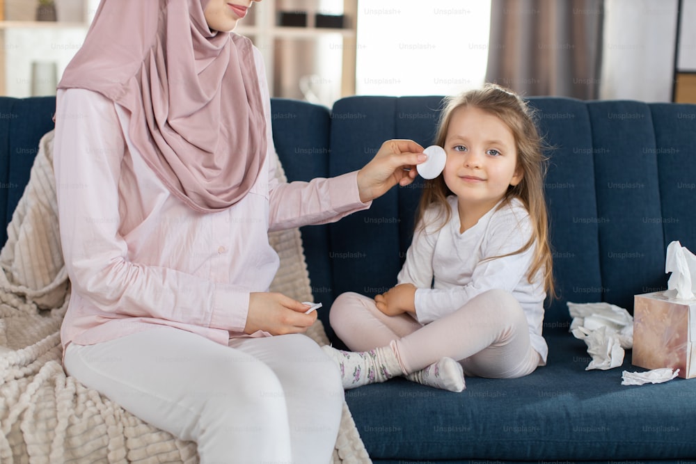Horizontal indoor shot of cute happy little girl kid sitting on blue sofa at home, while her muslim mother in hijab wiping and cleaning her cheek with cotton pad