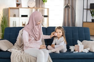 Happy family portrait. Children hygiene concept. Young muslim woman mother cleans with cotton pad face of her adorable pretty child girl, sitting on blue sofa at home