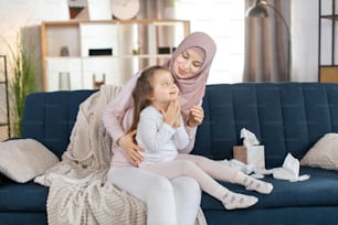 Baby hygiene and skin care concept. Cute little girl, applying moisturizing cream or baby lotion on her face, sitting on the knees of her young lovely Muslim mom in living room.