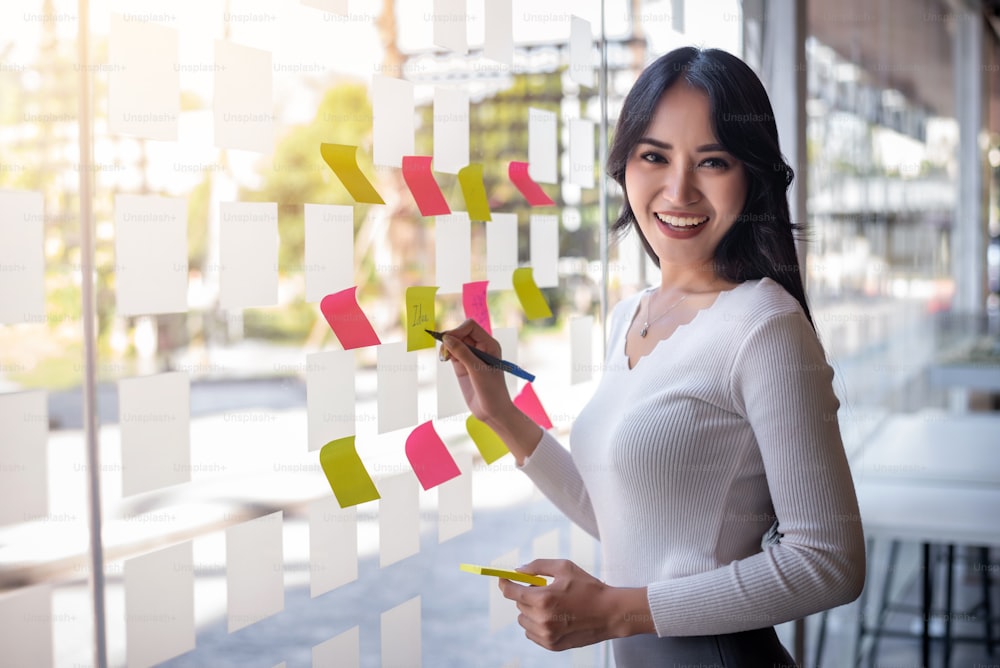 Portrait of young asian businesswoman beautiful charming smiling sticking sticky note on the glass wall in the office.