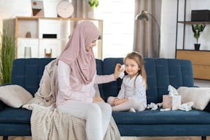 Front view of young pretty mother, Muslim woman in hijab, sitting on blue couch at home and wiping face of her cute little 3 years old daughter with paper napkin. Morning hygiene routine at home.