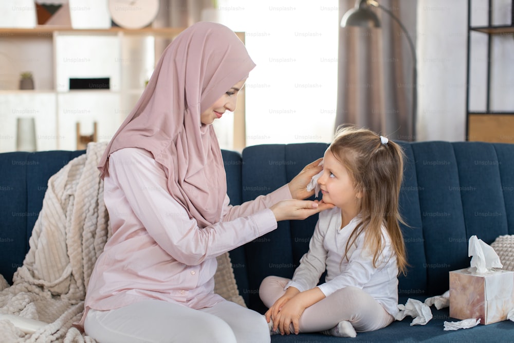 Young affectionate Muslim mother, wearing beige hijab, sitting on blue sofa at cozy home interior, and wiping face of her little happy daughter. Mom and child looking each other.