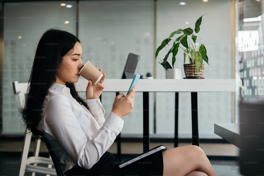 Des femmes d’affaires utilisent un téléphone intelligent pour lire et envoyer des messages texte tout en tenant une tasse de café à son bureau.