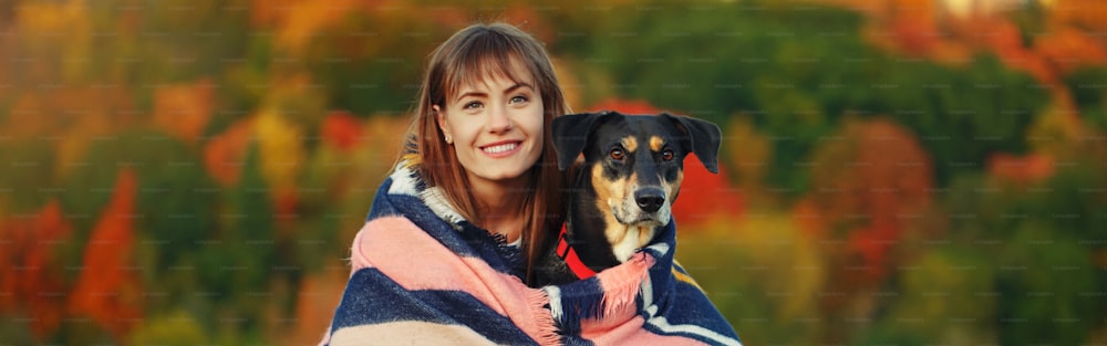 Donna avvolta in una coperta di lana con cane nel parco autunnale autunnale. Bella giovane donna caucasica felice che abbraccia l'animale domestico domestico. Migliori amici per sempre. Intestazione banner per sito web.