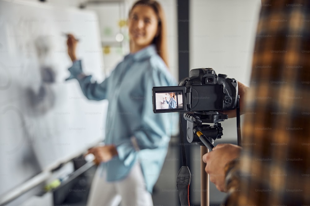 Beautiful smiling pleased dark-haired young Caucasian engineer being filmed by her colleague in the office