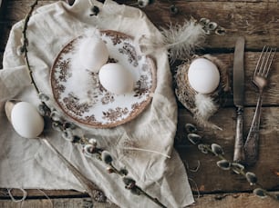 Stylish rustic Easter table setting. Natural easter egg in nest, vintage plate and cutlery, soft feathers, linen napkin, pussy willow branches on rustic wooden table. Rural Easter still life