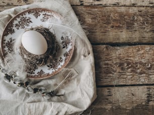 Uovo di Pasqua naturale in nido con morbide piume su piatto vintage, tovagliolo di lino, rami di salice figa su legno invecchiato. Elegante tavola di Pasqua rustica. Natura morta pasquale rurale. Buona Pasqua