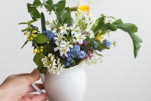 Hand hält schöne Frühlings-Wildblumen in Vintage-Tasse. Blühende bunte Blüten von Ajuga, Vergissmeinnicht, Narzissen, Pulmonaria, Rabelera holostea Nahaufnahme. Bäuerlicher Wiesenstrauß