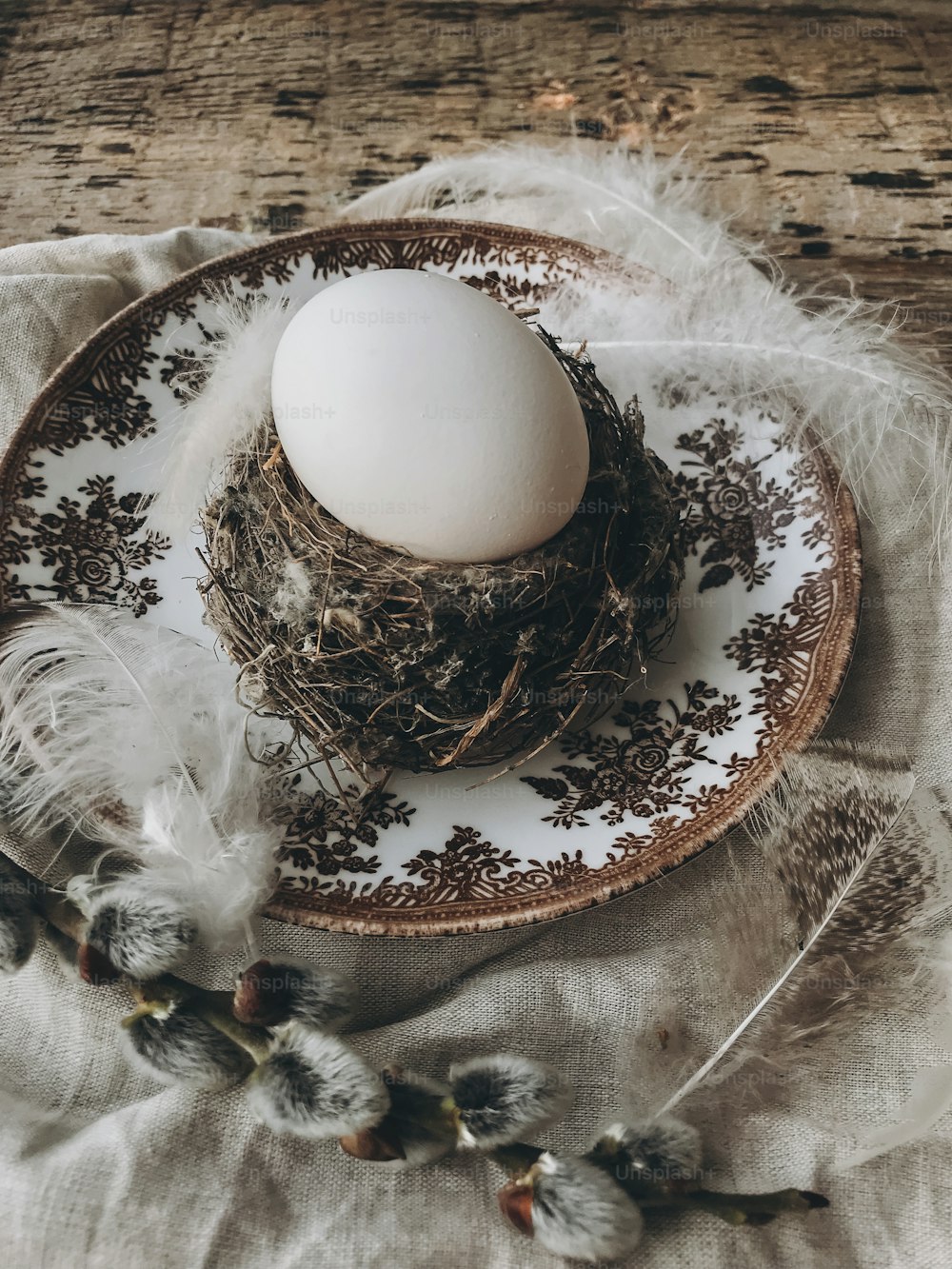 Stylish rustic Easter table setting. Natural easter egg in nest with soft feathers on vintage plate, linen napkin, pussy willow branches on aged wood. Rural Easter still life. Happy Easter