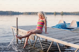 Funny happy cute Caucasian blonde girl child sitting on wooden deck pier by lake. Smiling laughing kid in swimsuit splashing with legs in water. Summer fun outdoor activity. Happy childhood lifestyle.