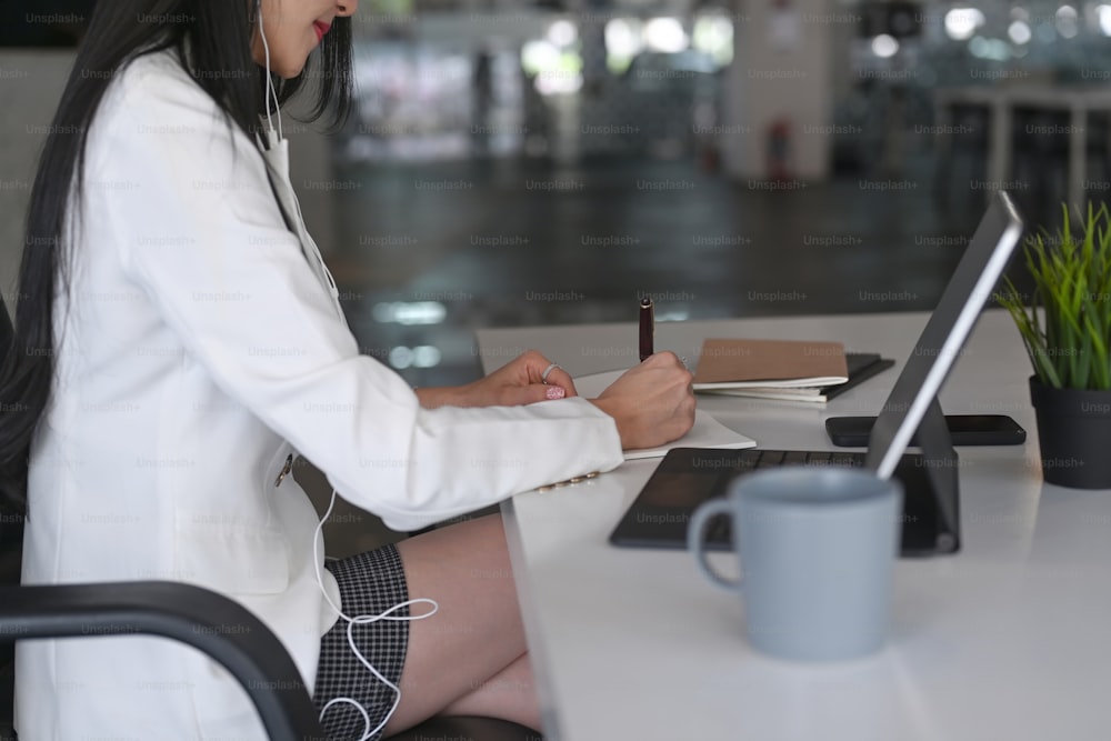 Side view of businesswoman wearing earphone and writing information on notebook at office desk.