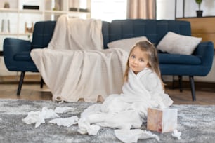 Portrait of beautiful little girl child wrapper in white towel after bath or shower, sitting on gray carpet in living room at home and smiling to camera. Paper napkins on the carpet. Copy space.
