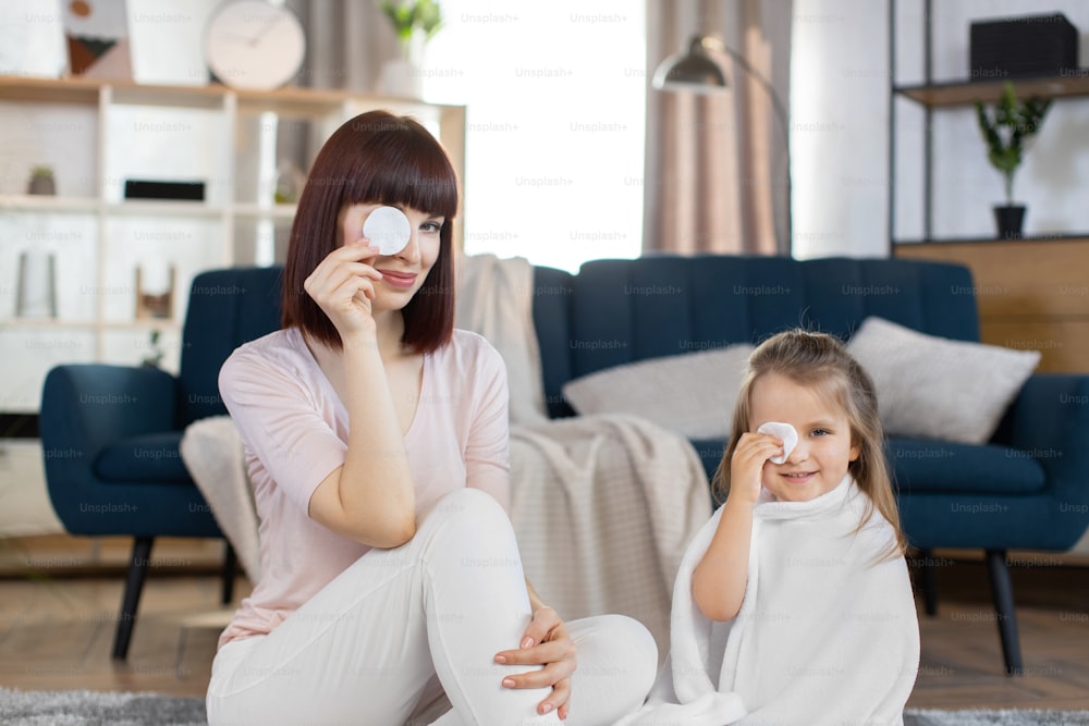 Beautiful smiling young woman mom, sitting on the floor at cozy room together with her little cute daughter wrapped in bath towel, and holding cotton pads on eyes. Family beauty spa concept.