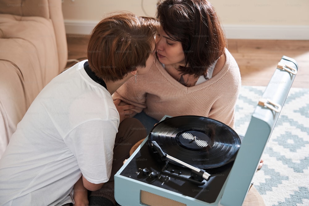 Happy lovely lesbian woman dressed in casual clothes listening music on vinyl records with her cute girlfriend in room indoors while sitting on floor together. Family spending time together at home concept