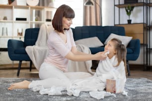 Young happy Caucasian woman mother, wiping with napkin face of her cute little 3 years old daughter, wrapped in a white towel after shower, sitting together at cozy room at home. Baby's hygiene