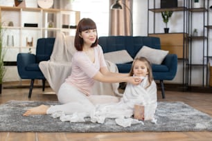 Young mom and little pretty girl child, sitting on the floor in living room at home. Cute girl is wrapped in towel after bath. Smiling mother wiping baby's face with napkin.