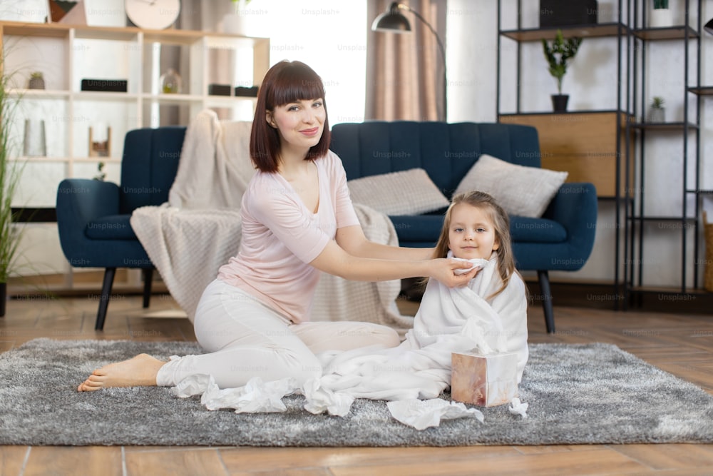 Mamá joven y niña bonita, sentada en el suelo en la sala de estar de casa. La chica linda está envuelta en una toalla después del baño. Madre sonriente limpiando la cara del bebé con una servilleta.