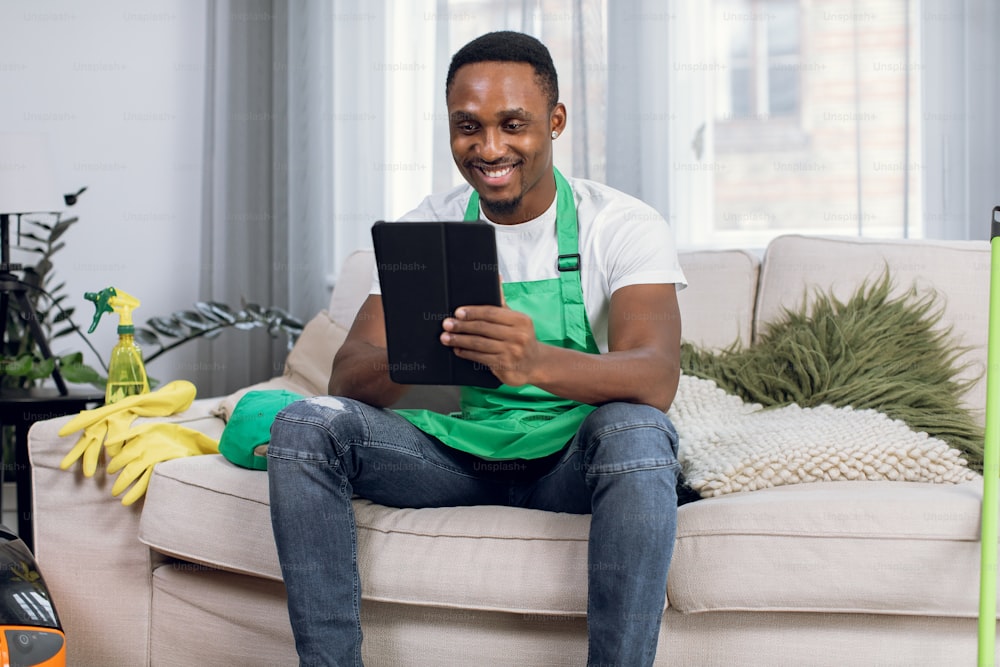 Close up of handsome african man cleaner in green apron using digital tablet while relaxing on couch after work. Male janitor sitting on sofa with modern cleaning equipment around.