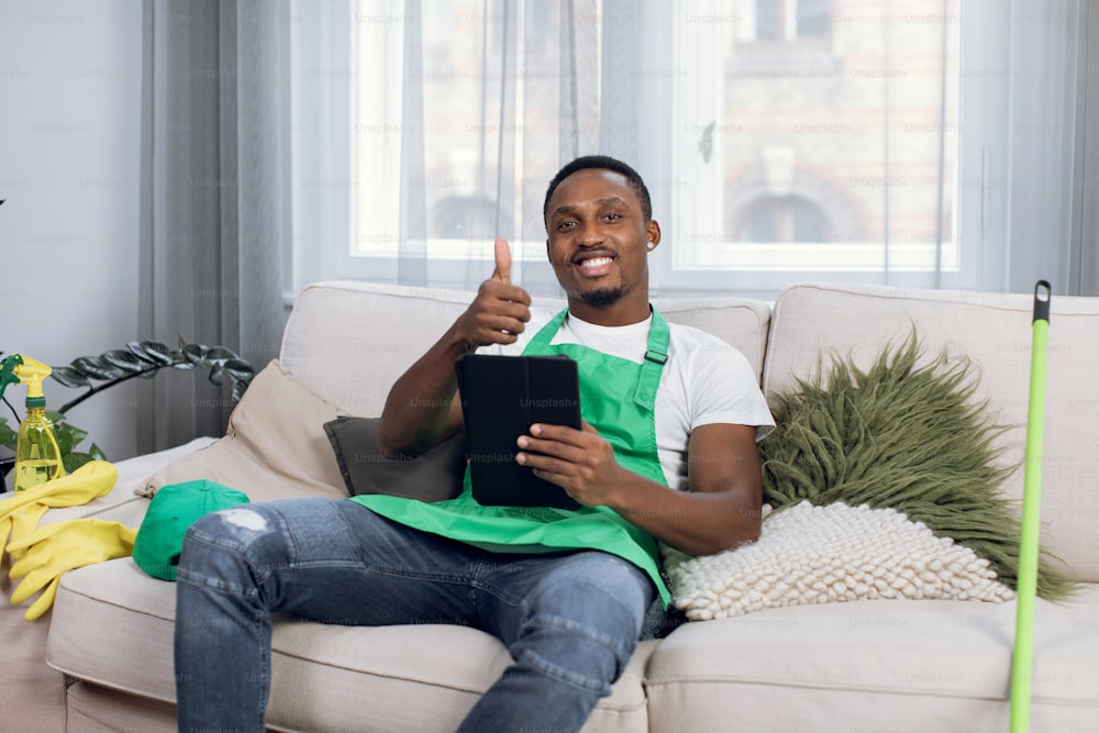 Smiling afro-american man in uniform holding digital tablet in hands while sitting on couch and shows thumb up to camera. Male janitor relaxing on sofa during cleaning process at modern apartment.