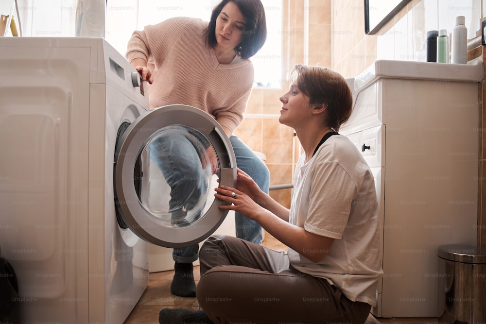 Couple doing laundry together putting clothes to the washing machine. Couple in a laundry room washing clothes with window in the background. LGBT lesbian couple love moments and happiness concept