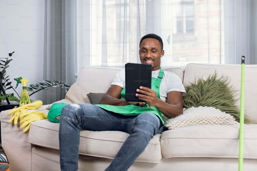Uomo afroamericano sorridente in uniforme che tiene la tavoletta digitale in mano mentre è seduto sul divano. Custode maschio che si rilassa su un comodo divano durante il processo di pulizia in un appartamento moderno.