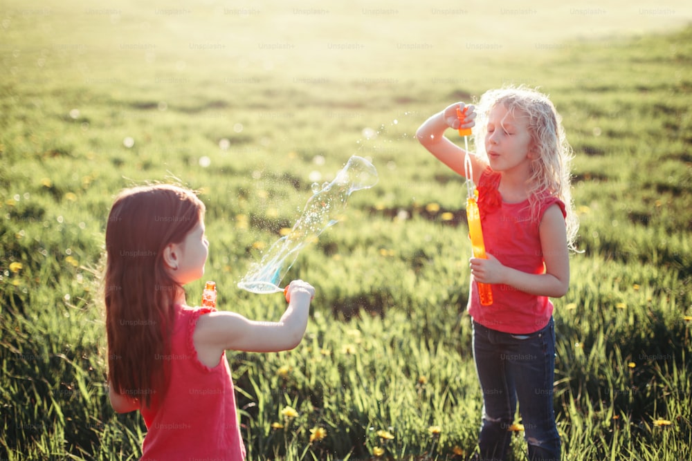 Preschool Caucasian girls blowing soap bubbles in park on summer day. Kids having fun outdoor. Authentic happy childhood magic moment. Lifestyle seasonal activity for children.