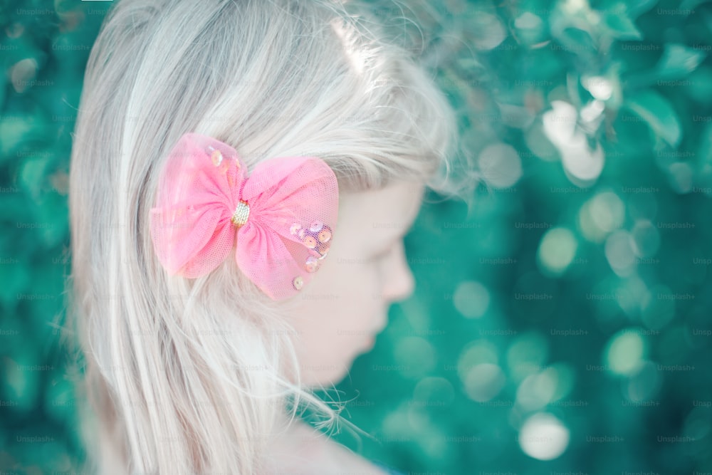 Portrait of little blonde Caucasian girl with pink hair bow. Pretty pensive sad child kid. Girly girl with red hair clip looking away. Lonely child. Soft selective focus. Blurry background.
