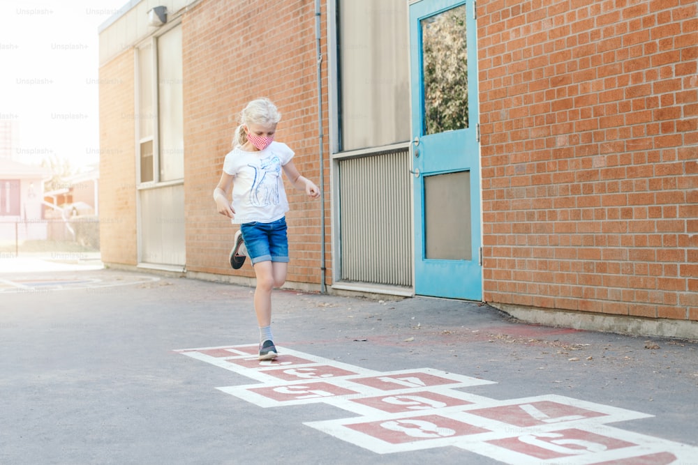 Child girl in face mask playing jumping hopscotch on school yard. Funny activity game for kids on playground outdoor. Street sport for children. Coronavirus covid-19 safety measures. New normal.