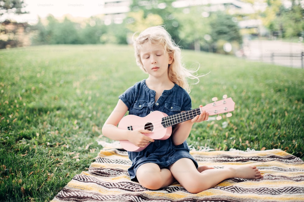 Cute adorable blonde girl playing pink guitar toy outdoor. Child playing music and singing song in park. Hobby activity for children kids. Tender memorable authentic candid childhood moment.