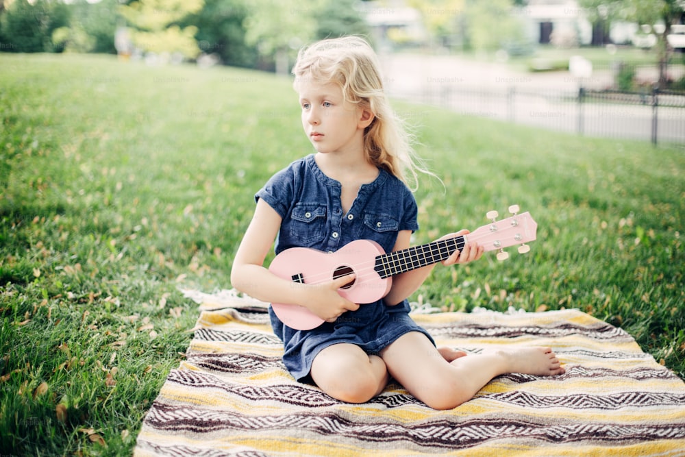 Cute adorable blonde girl playing pink guitar toy outdoor. Child playing music and singing song in park. Hobby activity for children kids. Tender memorable authentic candid childhood moment.