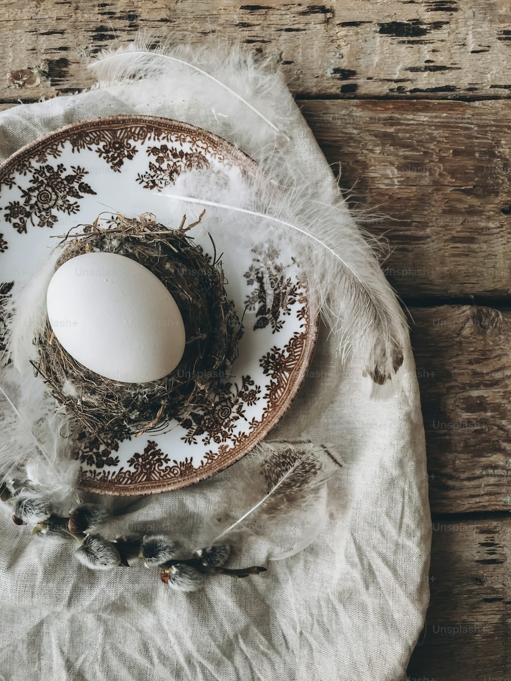 Natural easter egg in nest with soft feathers on vintage plate, linen napkin, pussy willow branches on aged wood. Stylish rustic Easter table setting. Rural Easter still life. Happy Easter