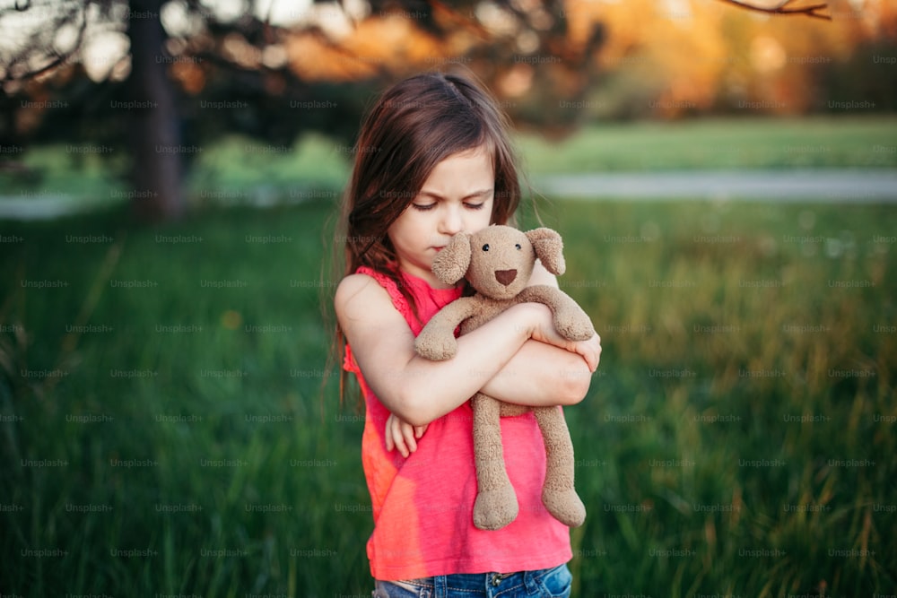 Sad upset Caucasian girl hugging toy. Child embrace soft plush bear in park outdoors. Lost lonely child kid outside. Unhappy childhood problems lifestyle.